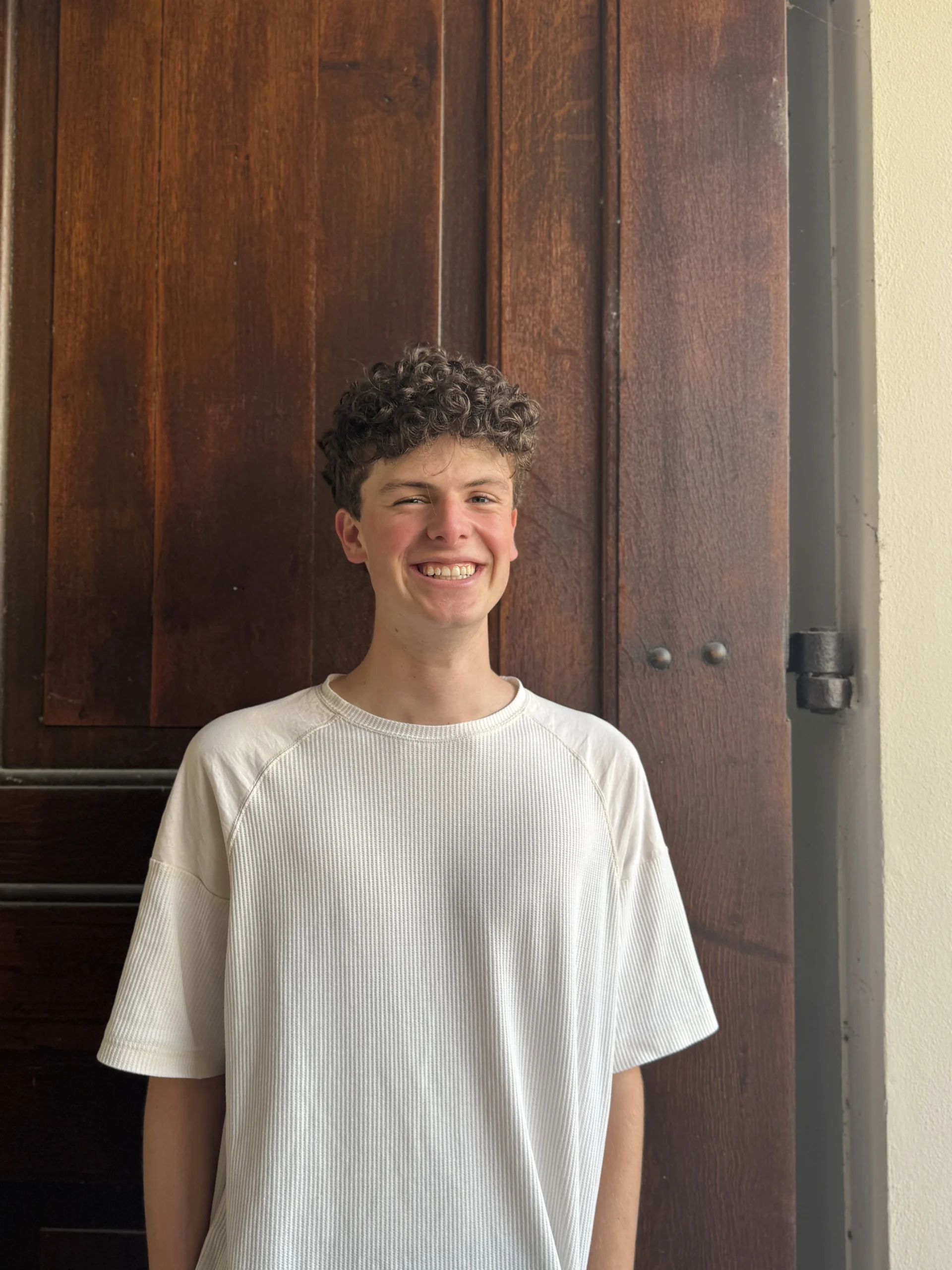 Junger Mann mit lockigem Haar in einem weißen T-Shirt steht lächelnd vor einer dunklen Holztür